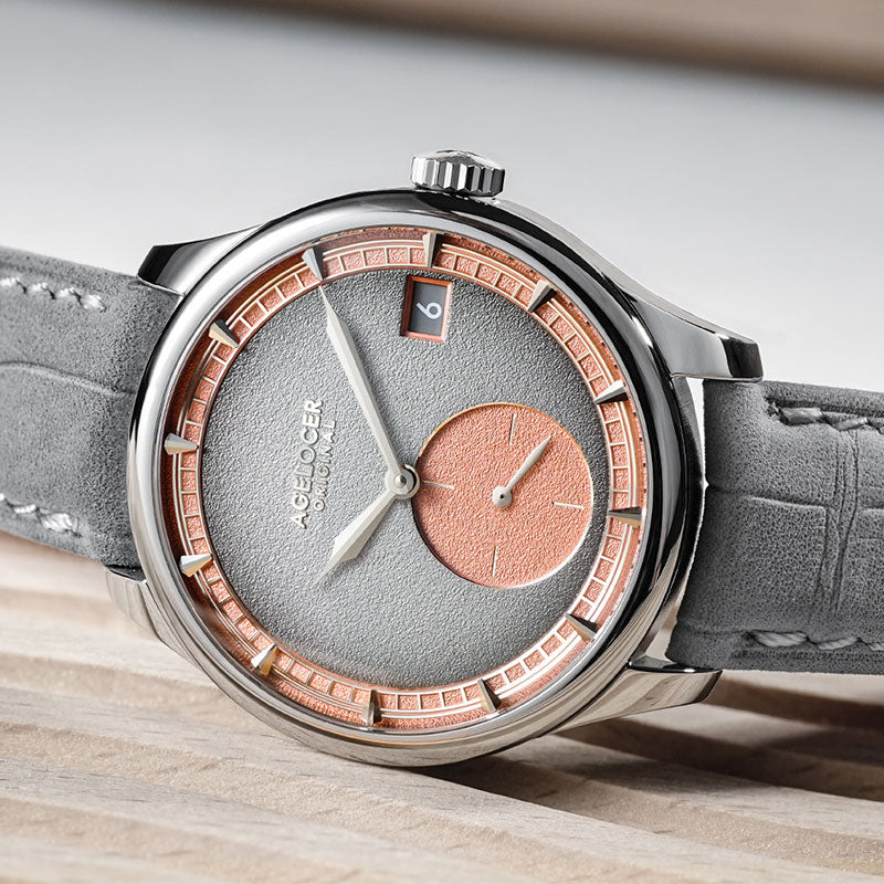 Agelocer Petite Seconde automatic watch with grey and salmon dial on a grey suede strap, resting on a wooden surface