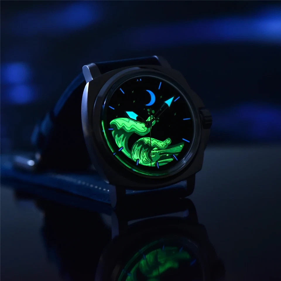 Luminous night view of Pagani Design H35A automatic mechanical watch with glowing blue dial, silver case and dark blue suede strap, photographed in a dimly lit setting.