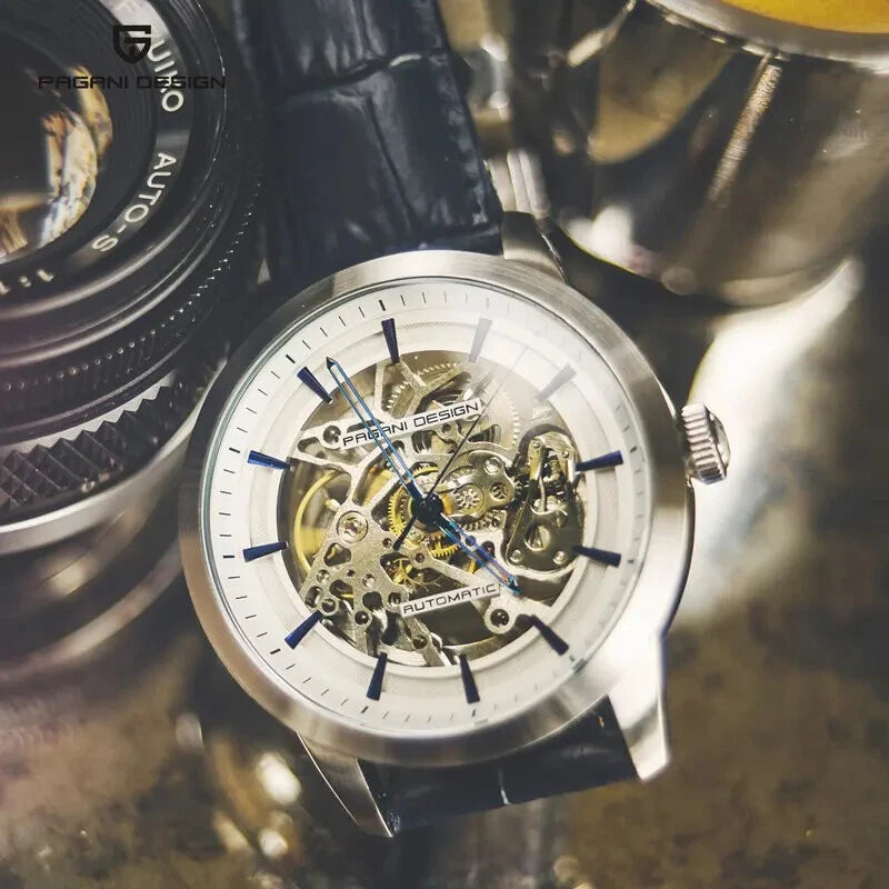 Lifestyle closeup of Pagani Design PD-1638 skeleton automatic watch with silver case, skeletonized dial, and blue leather strap, placed on a reflective surface with a camera and cup in the background.
