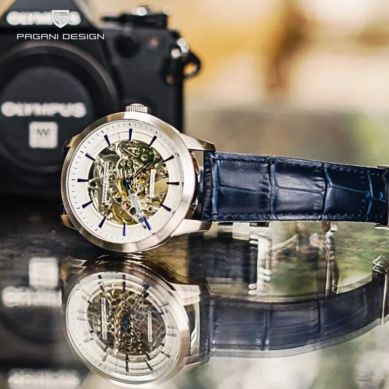 Lifestyle shot of Pagani Design PD-1638 skeleton automatic watch with silver case, skeletonized dial, and blue leather strap placed horizontally next to a camera with its reflection visible.