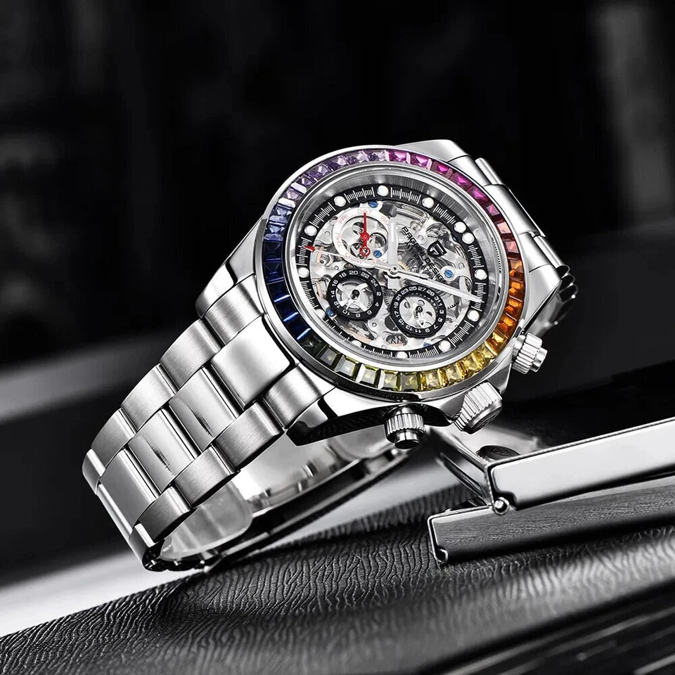Lifestyle shot of Pagani Design PD-1653 automatic mechanical chronograph watch with skeleton dial, rainbow bezel, and polished stainless steel bracelet resting on a black leather surface next to a silver cufflink with orange accent.
