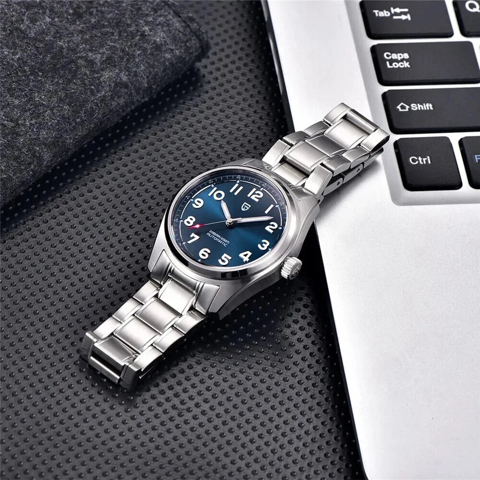 Lifestyle photo of Pagani Design PD-1717 automatic mechanical watch with blue dial and stainless steel bracelet resting on a black textured desk next to a laptop keyboard.