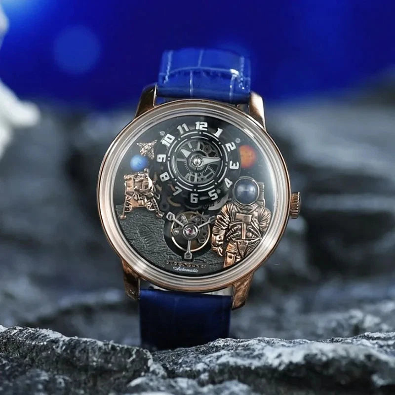 Pindu W2101B Space Series mechanical watch with three-dimensional astronaut and planet motif dial, rose gold case, blue leather strap, photographed on a rocky surface with blurred blue background.