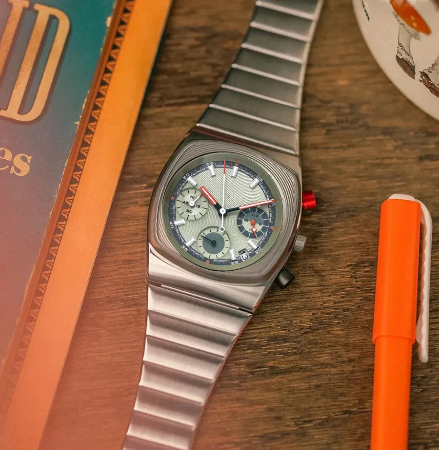 Silver wristwatch with a detailed face on a wooden surface next to an orange pen and book.