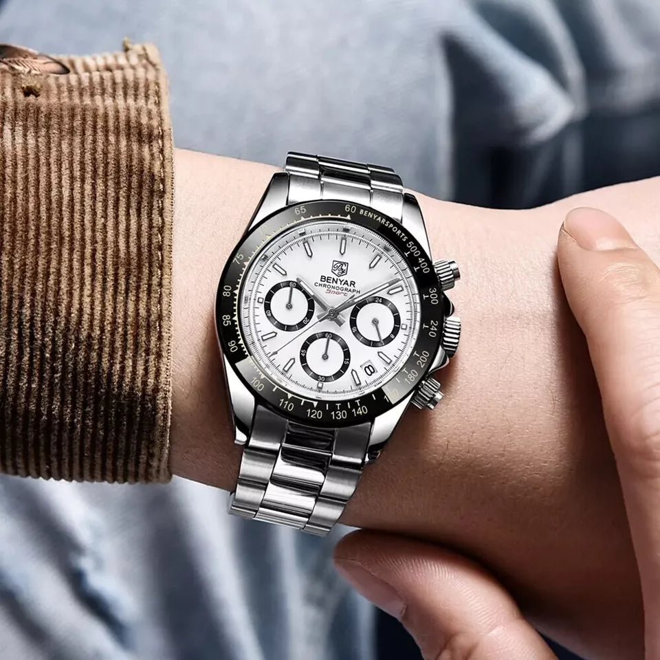 Silver wristwatch with a detailed face on a person's wrist, wearing a brown sleeve.