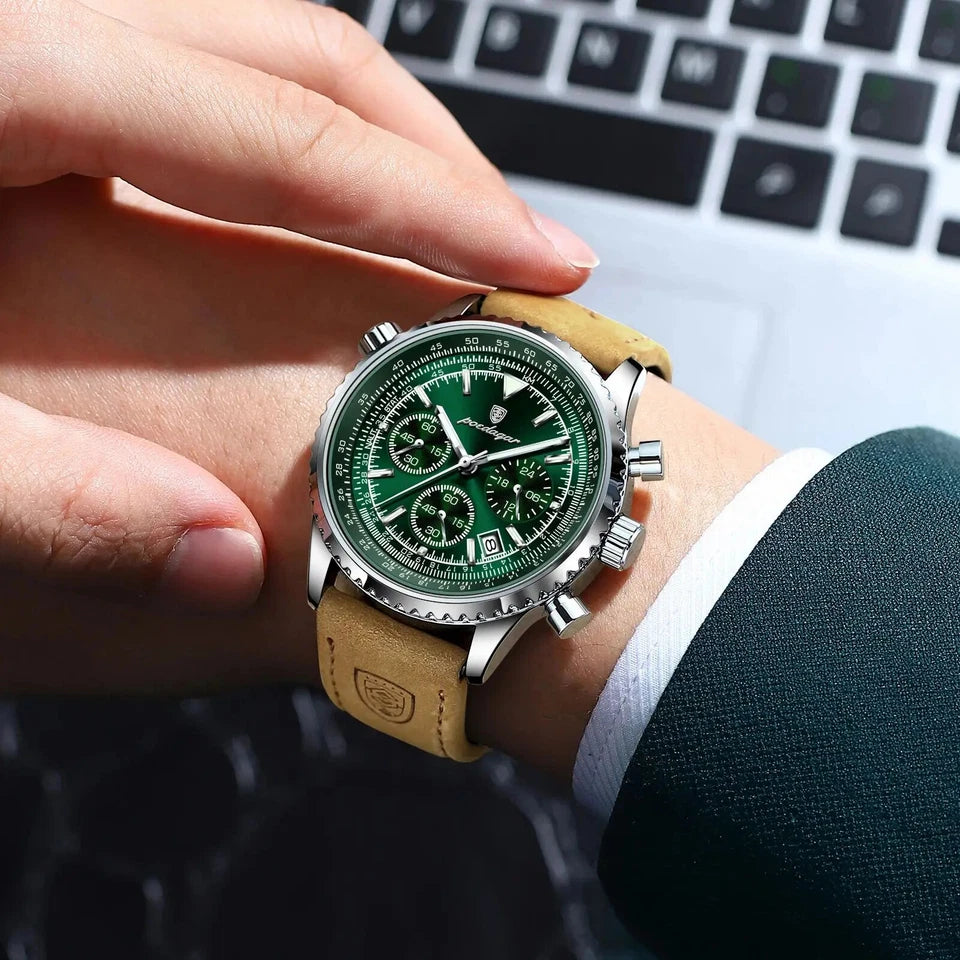 Green watch with brown leather strap on a person's wrist, blurred laptop in the background