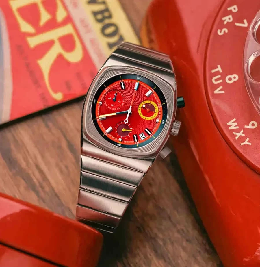 Silver watch with a red face on a wooden surface with a red phone and book in the background