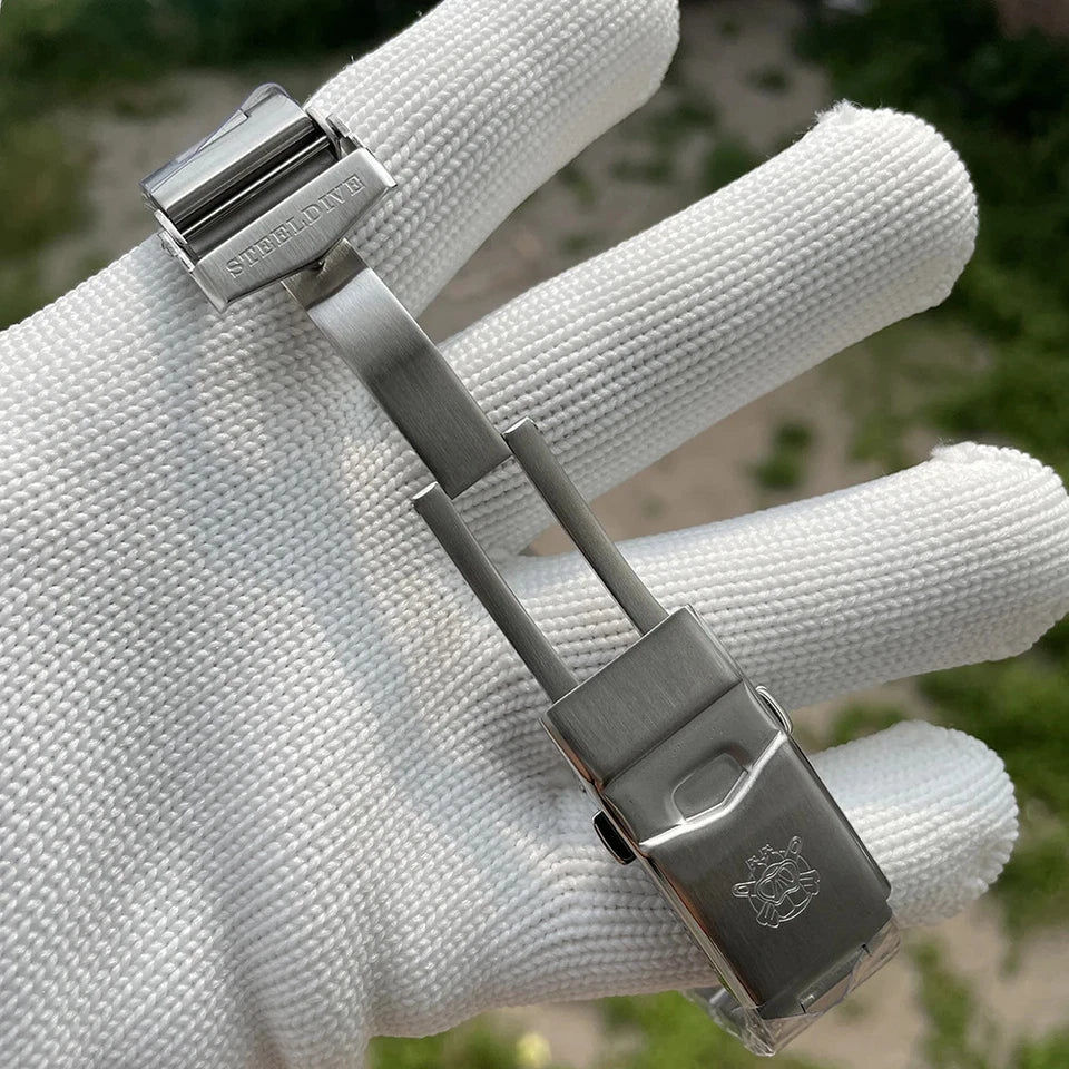 Steeldive SD1953 stainless steel watch strap buckle and clasp displayed on a white glove with blurred green background.