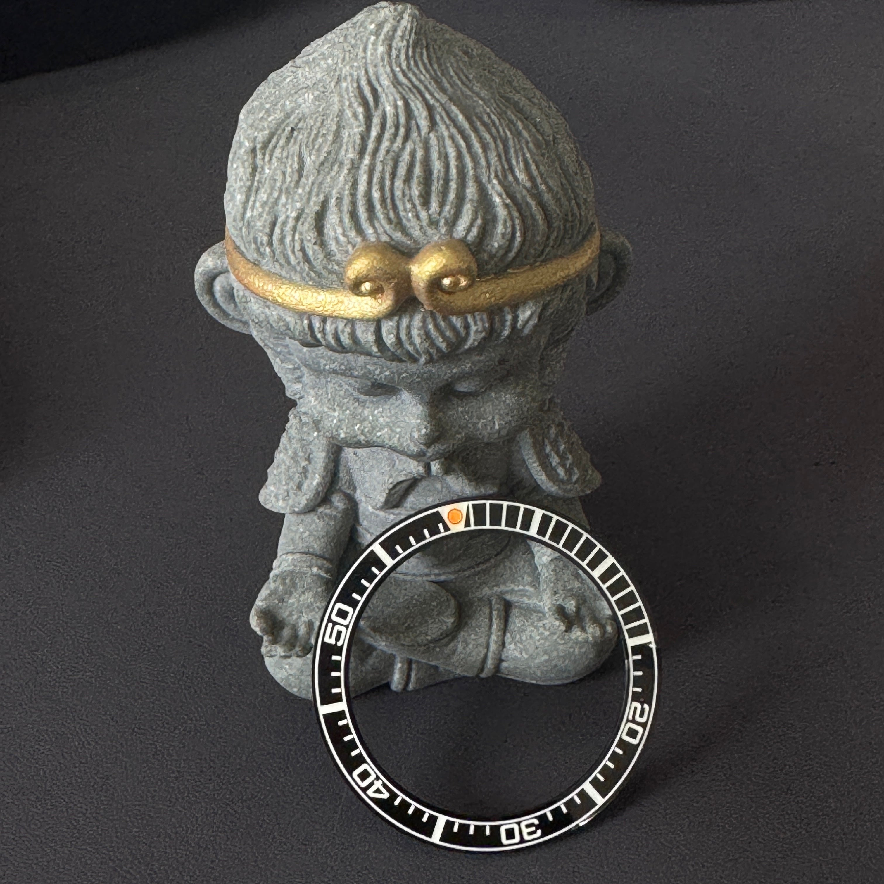 Steeldive SD1961 ceramic bezel insert leaning on stone figurine, white 60-minute scale and orange pip visible