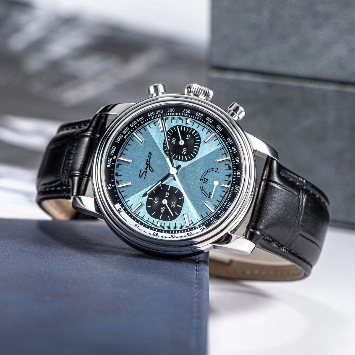 Sugess S438 ice blue chronograph, black sub-dials, black leather strap, angled view beside grey box