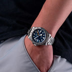 Sugess S463 GMT blue dial watch worn on the wrist over grey trousers, steel bracelet